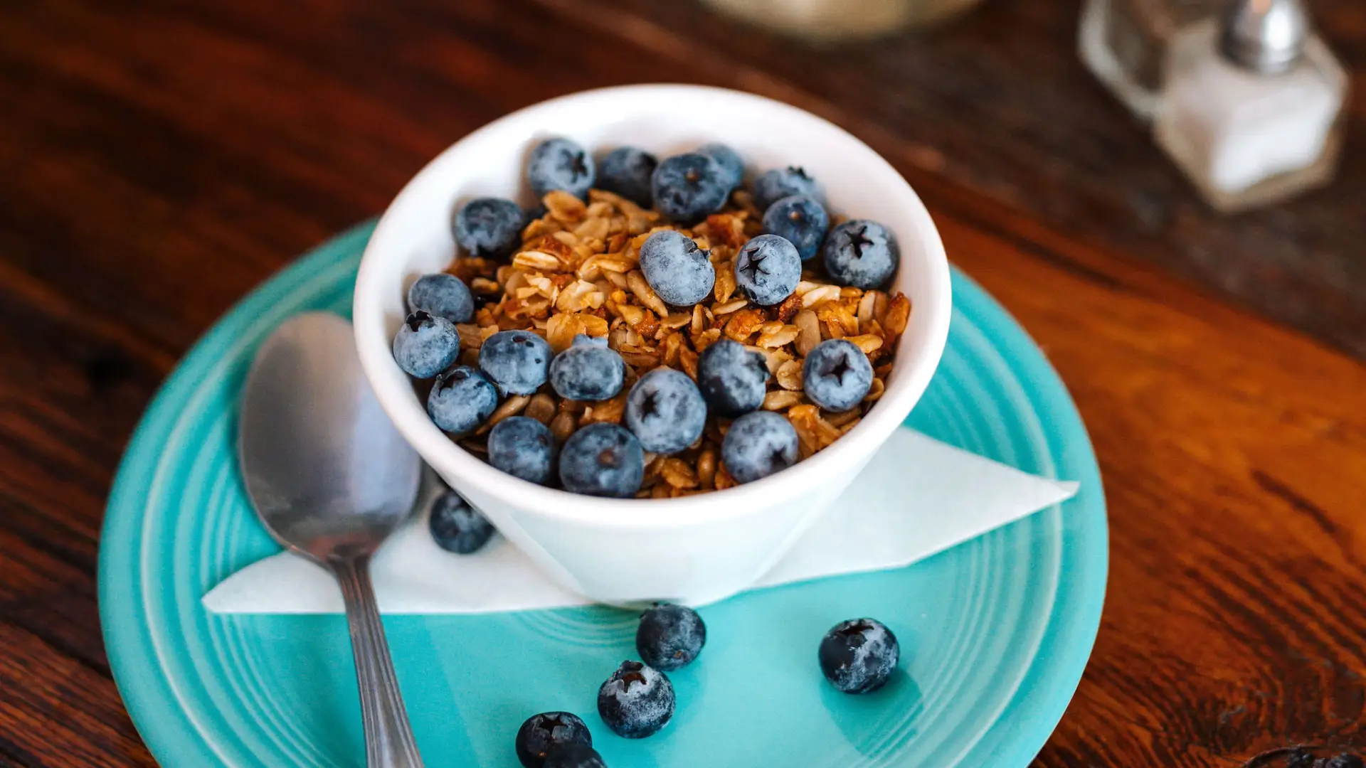 Berries and granola overflow their bowl onto the blue plate below the yogurt bowl.