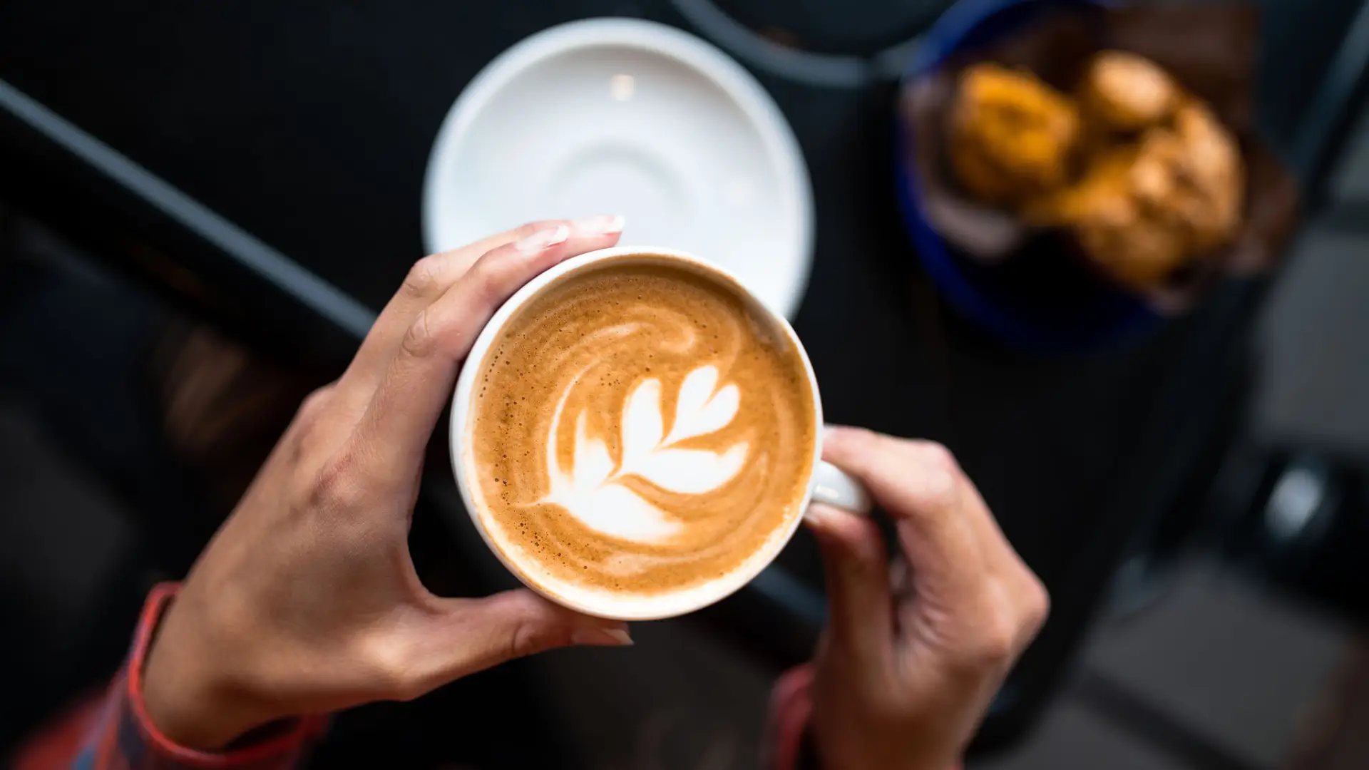 Hands holding a latte with art over a white saucer.