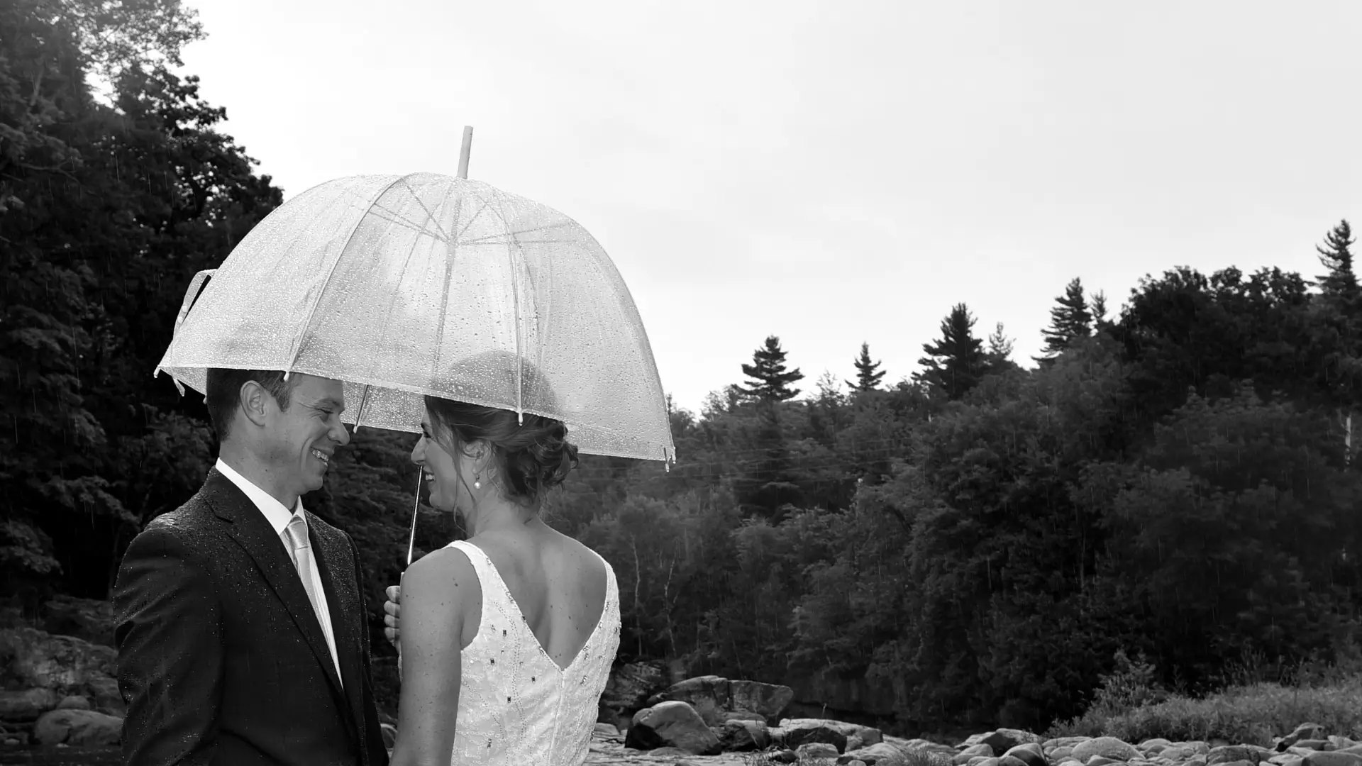 A bride and groom stand by a riverbed under a clear umbrella in black and white.