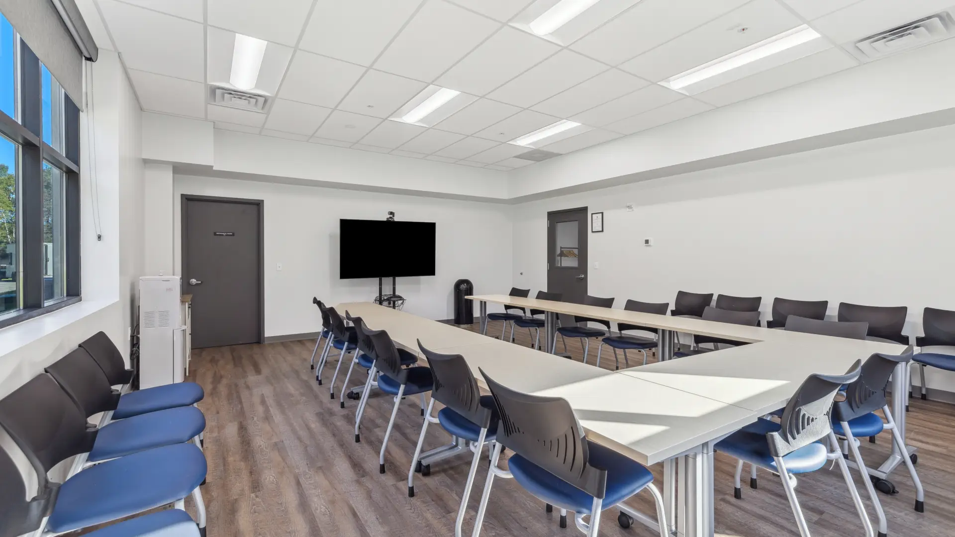 The meeting space arranged in a U shape with chairs and a large screen on a mount, viewed from the corner opposite the door.