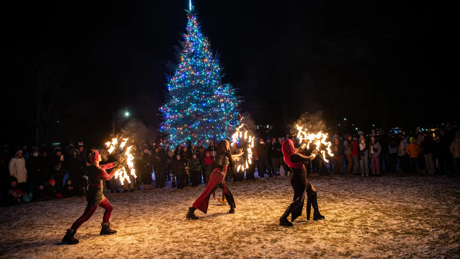 Three fire dancers perform for a crowd in front of a large lit up Christmas tree in winter. 