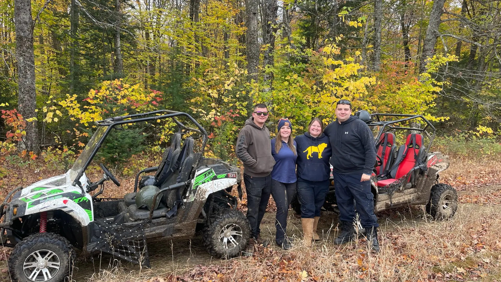 Four people stand together between two muddy UTVs surrounded by fallen leaves and vibrant fall foliage 