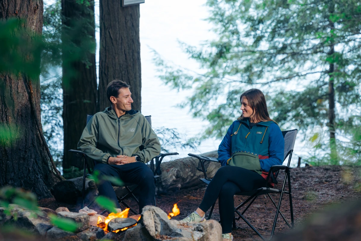 A man and woman sit in camp chairs at a campsite on a lake. 