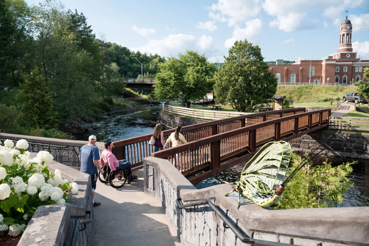 People, including one in a wheelchair, cross a pedestrian bridge surrounded by flowers in a charming downtown.