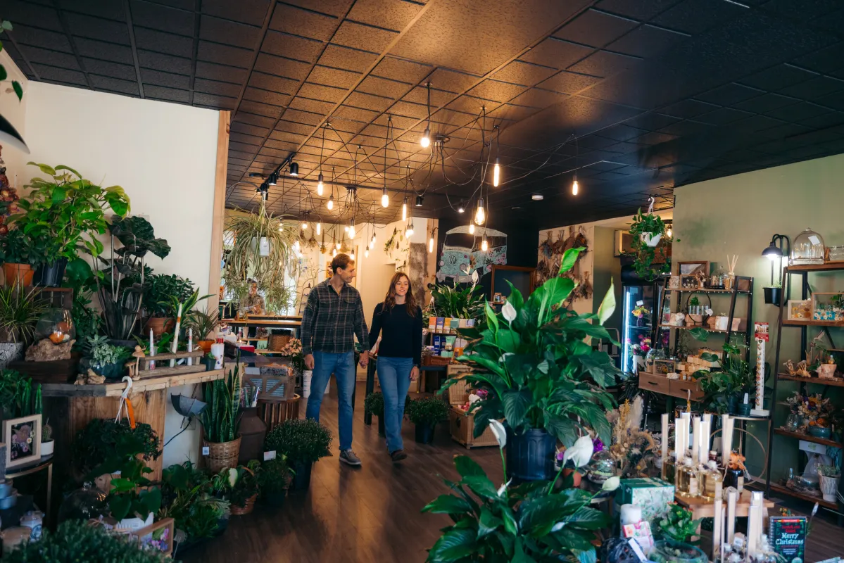 A man and woman walk through a greenhouse shop filled with plants. 