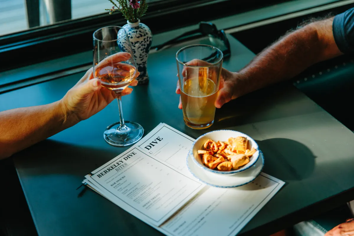 Two adults hold glasses of alcohol over a menu and bowl of snacks at a restaurant.