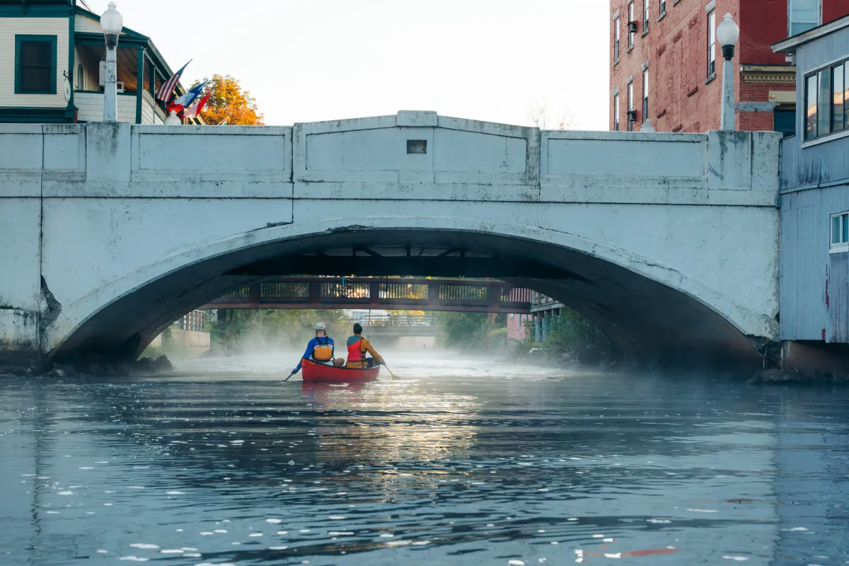 Two people paddle in a canoe on a river flowing under a bridge.