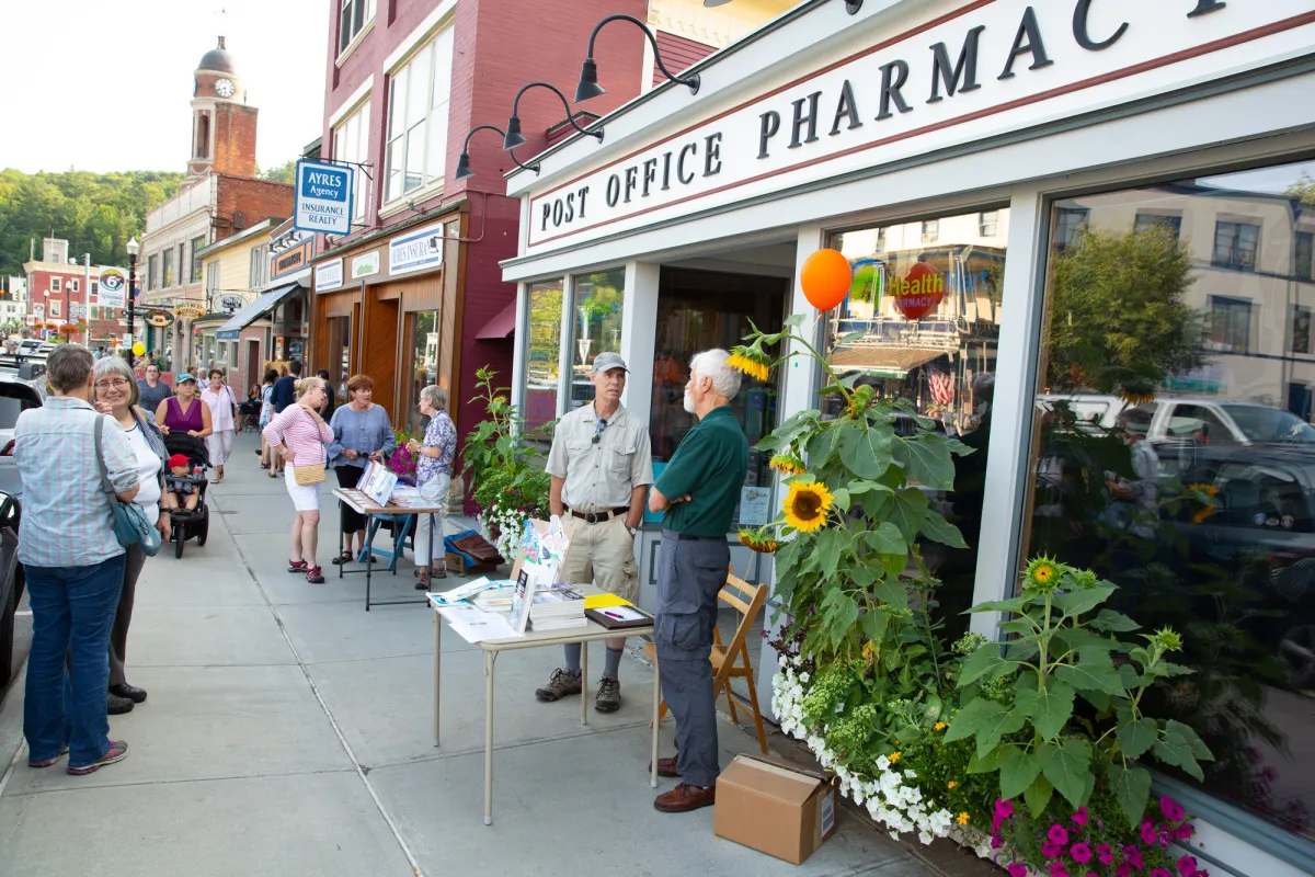 A pharmacy and post office on a main street with people walking the sidewalk.