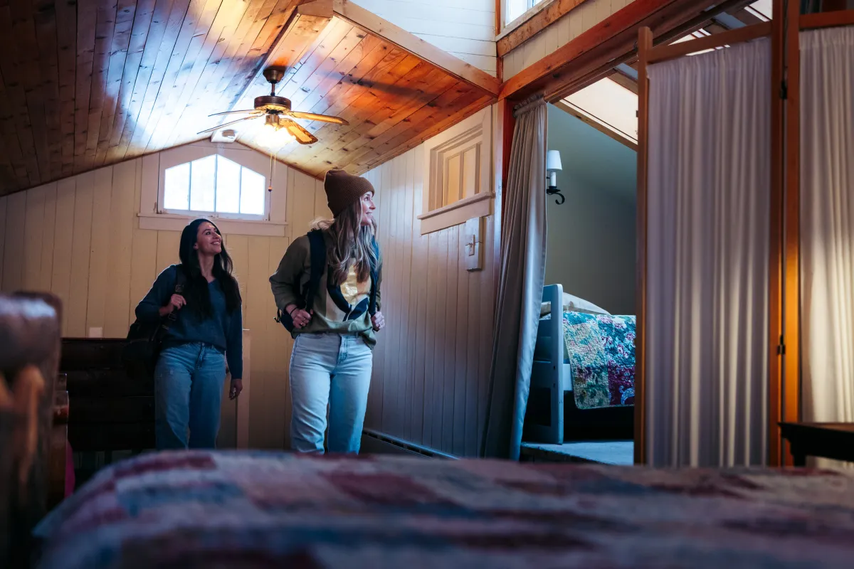 Two women walk into a bedroom of a rustic home. 