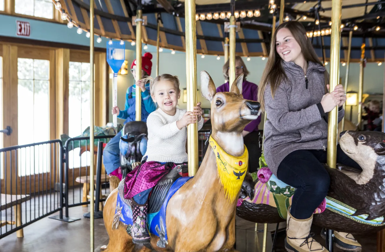 A woman laughs and rides a carousel with her young daughter.