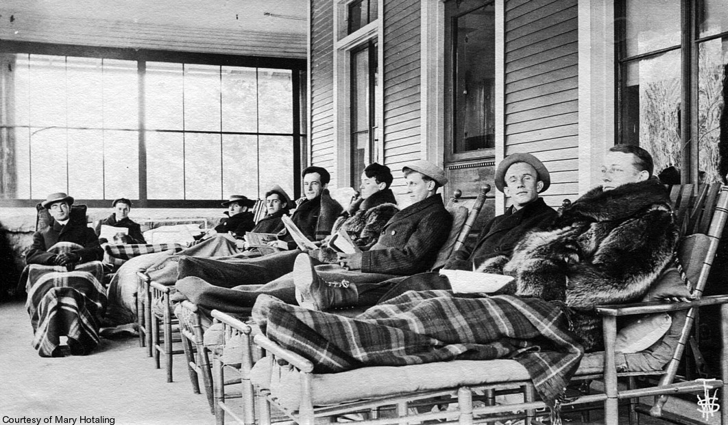 In a black and white image from the 1920s, male patients rest on lounge chairs on a sanitarium porch.