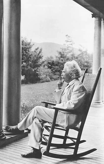 A black and white image of Mark Twain sitting in a rocking chair on a porch with a forest view in the background.