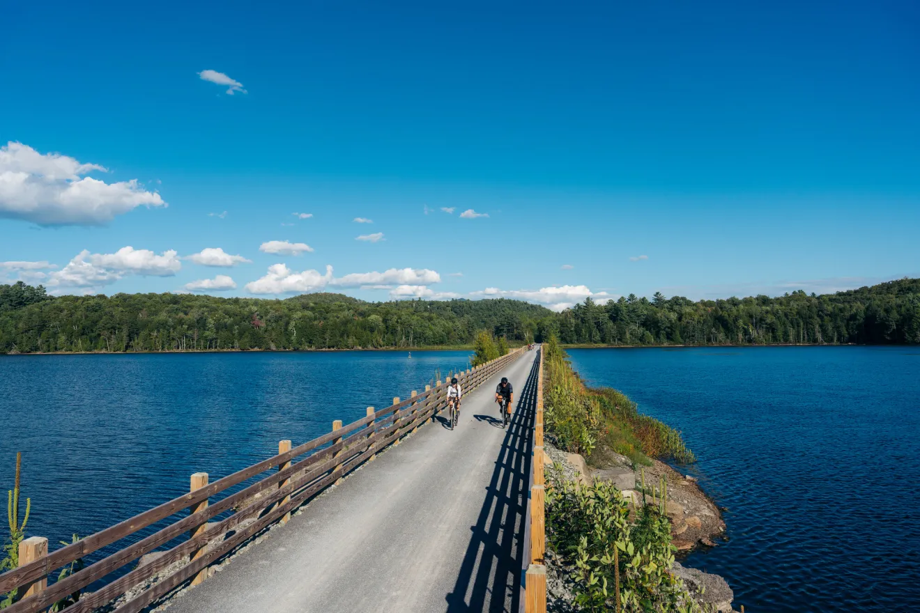 Two cyclists on a gravel pathway with lake and forested shorelines on either side.