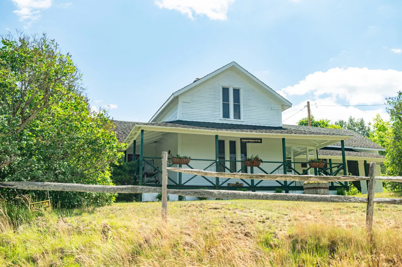 A white cottage with green trim sits atop a small slope of green grass on a sunny summer day.