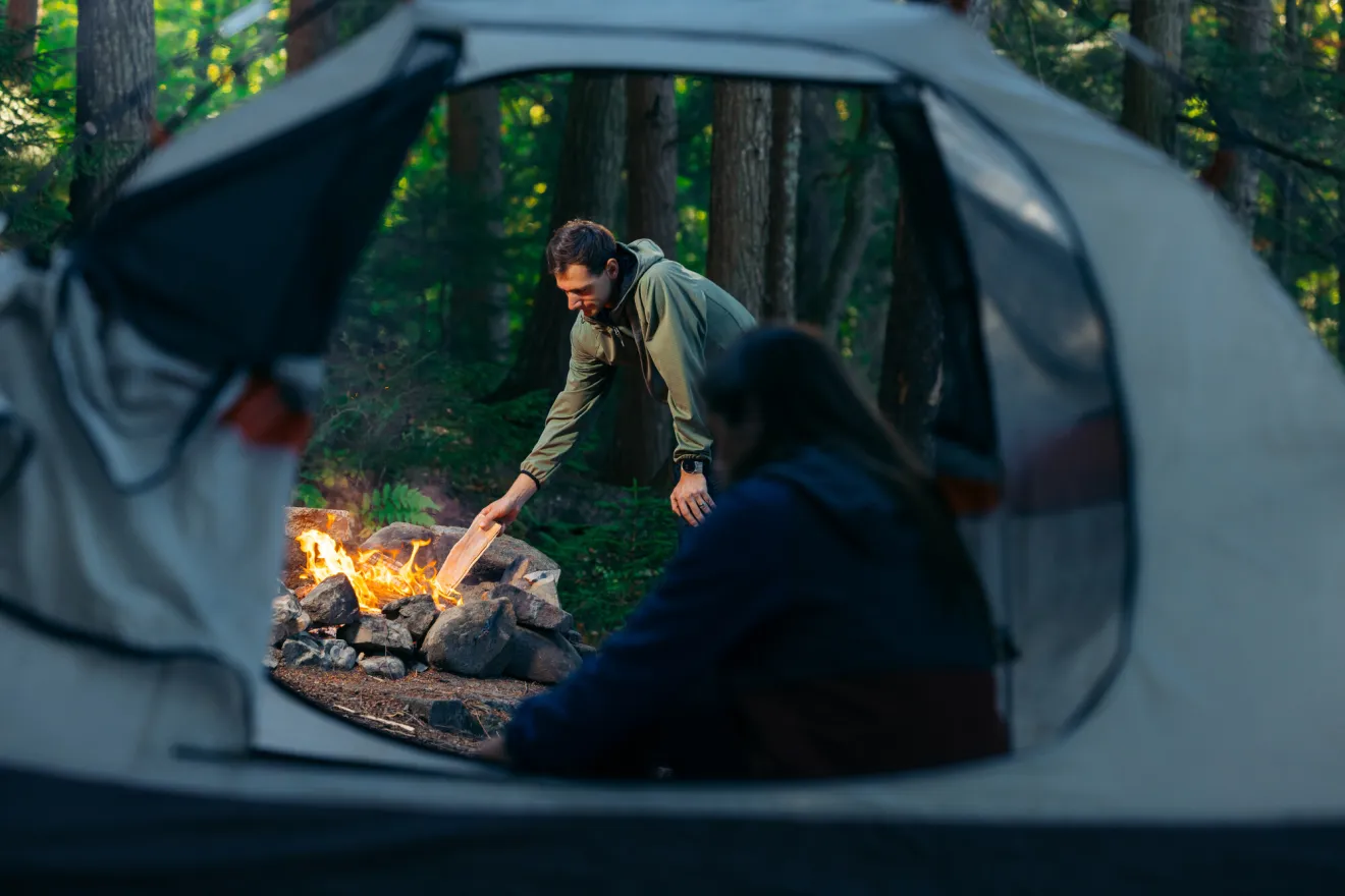 A woman watches a man stoke a fire from her tent. 