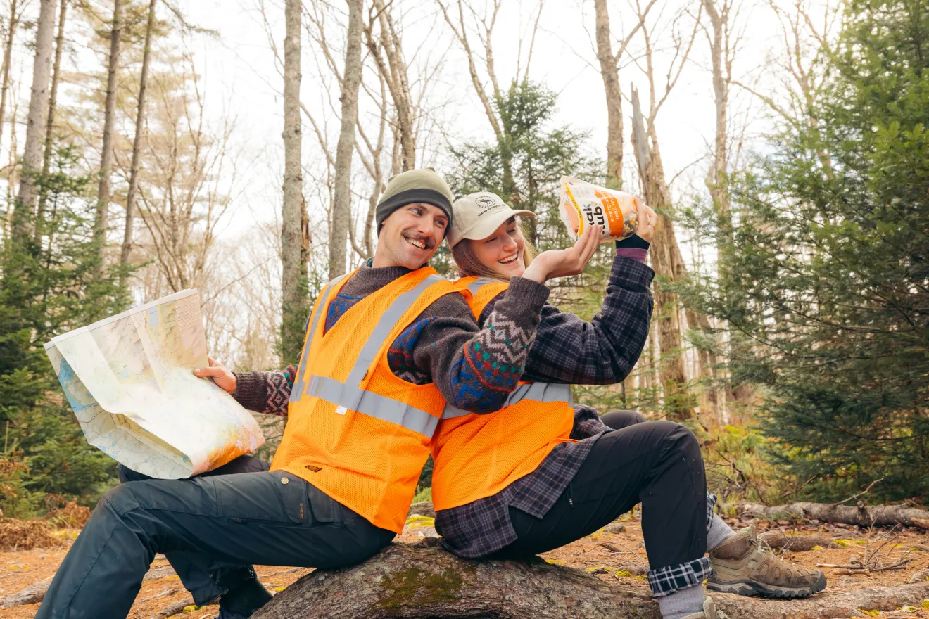 A man and a woman wear blaze orange vests in the woods. 