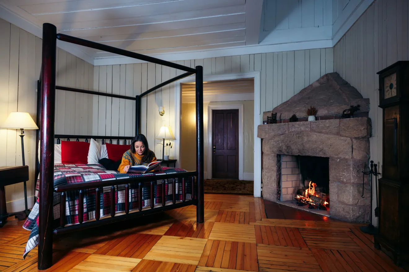 A woman lays on a bed reading a book in a cabin with a fire roaring.
