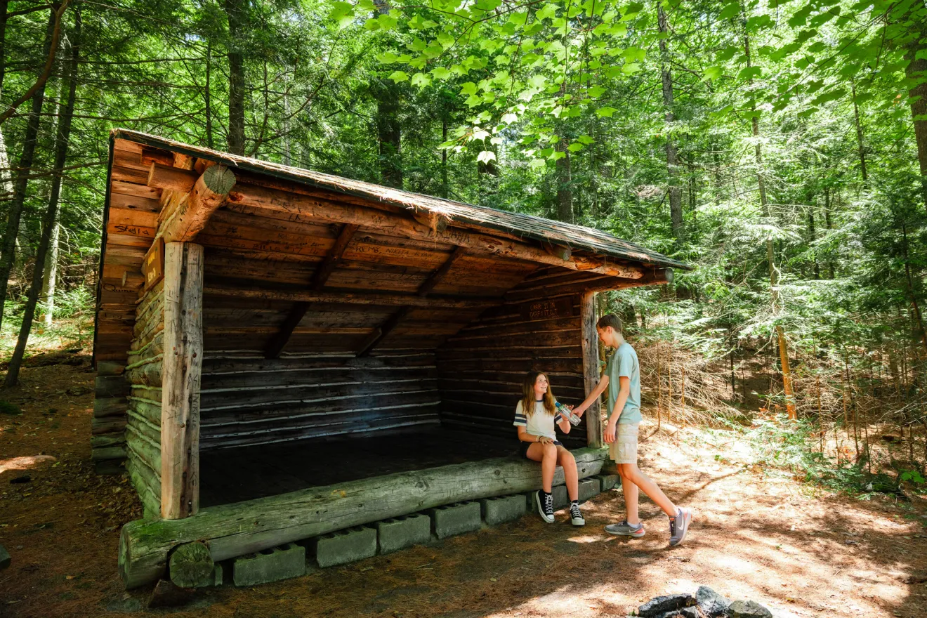 A young man and woman sit in a lean to on a hiking trail. 