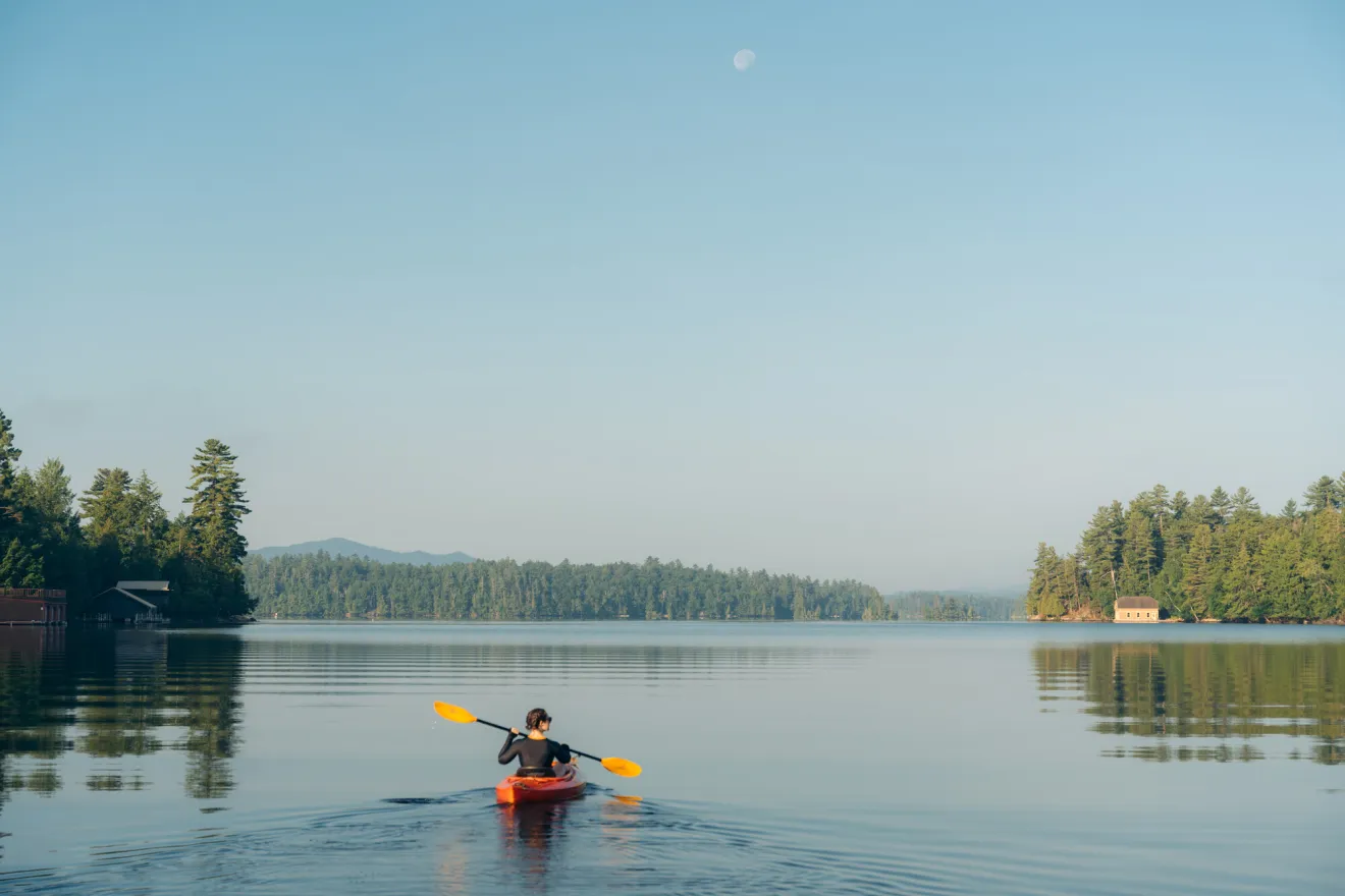 A solo female paddles a red kayak across a calm, flat pond. Treed shorelines are in the distance.