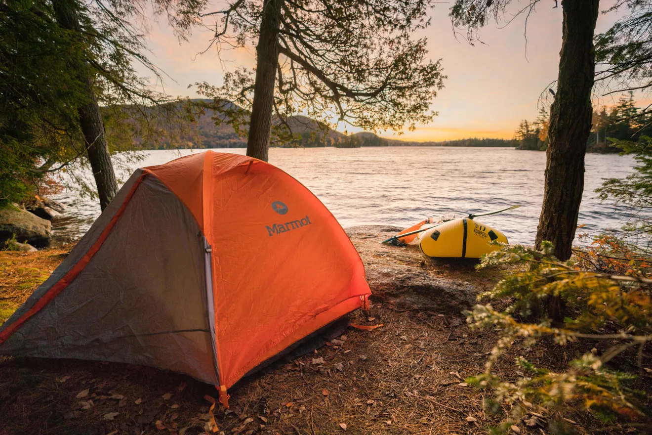An orange tent sits on the shore of a lake with a yellow raft laying next to it. 