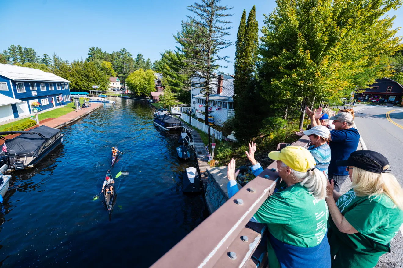 A group of people stand on a bridge and cheer as people paddle in canoes below. 