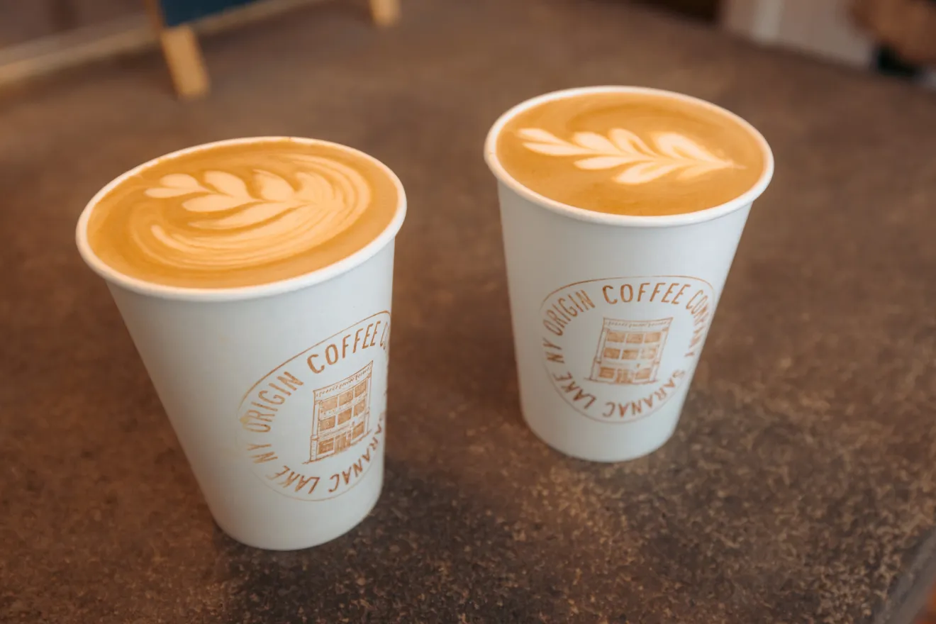 A close-up view of two paper cups of creamy coffee, a cream leaf pattern on the surface of the drink.