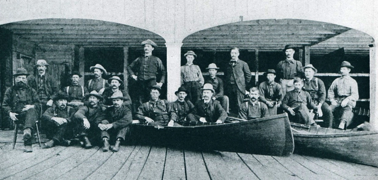 A group of outdoor guides at a large boathouse in a black and white image from the late 1800s.