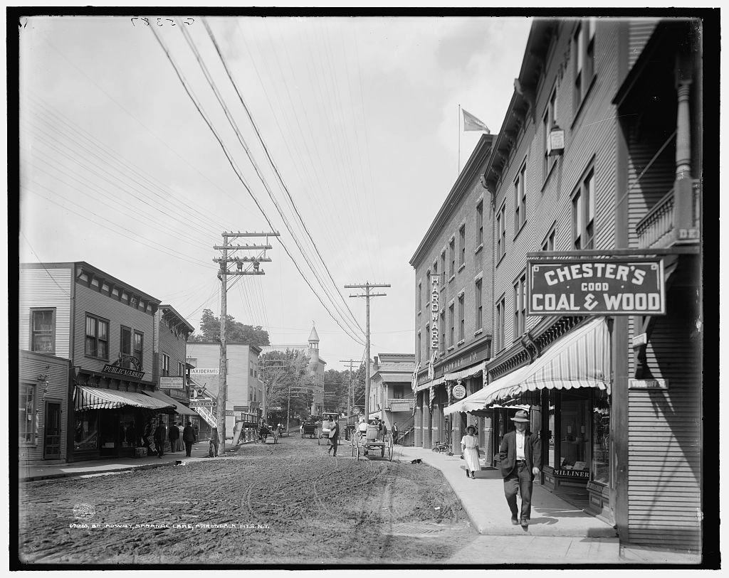 A black and white image of a muddy main street in a small town, ca. 1909. Shops line the street.
