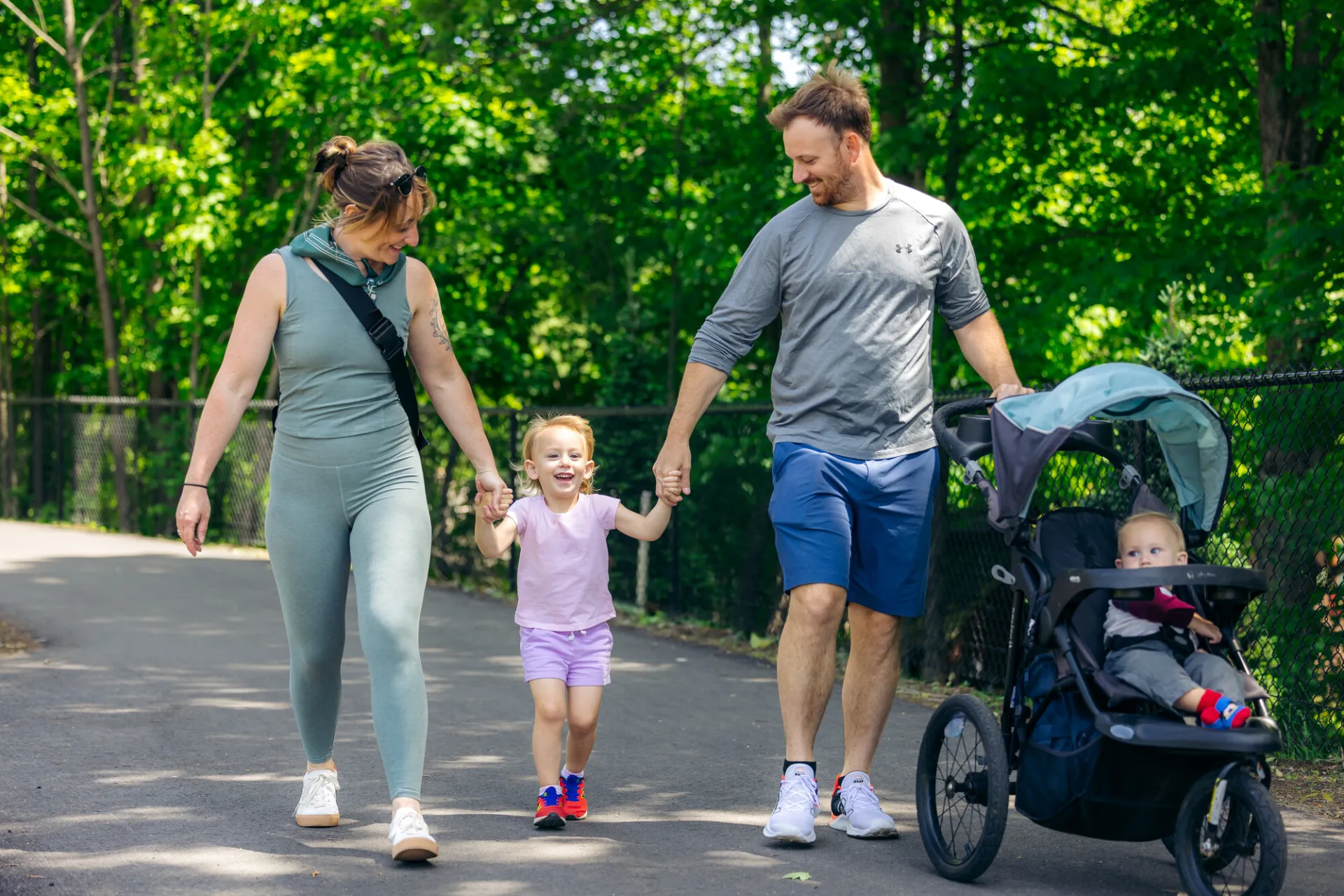 A man and woman walk a young girl and stroller on a recreation trail.