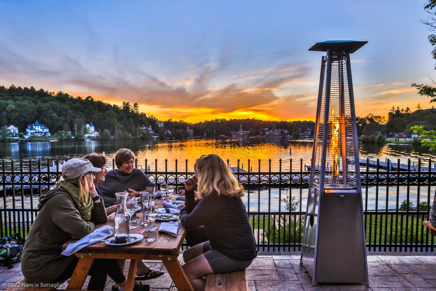 Women gather at a picnic table on a restaurant patio, overlooking a calm lake and a sunset.