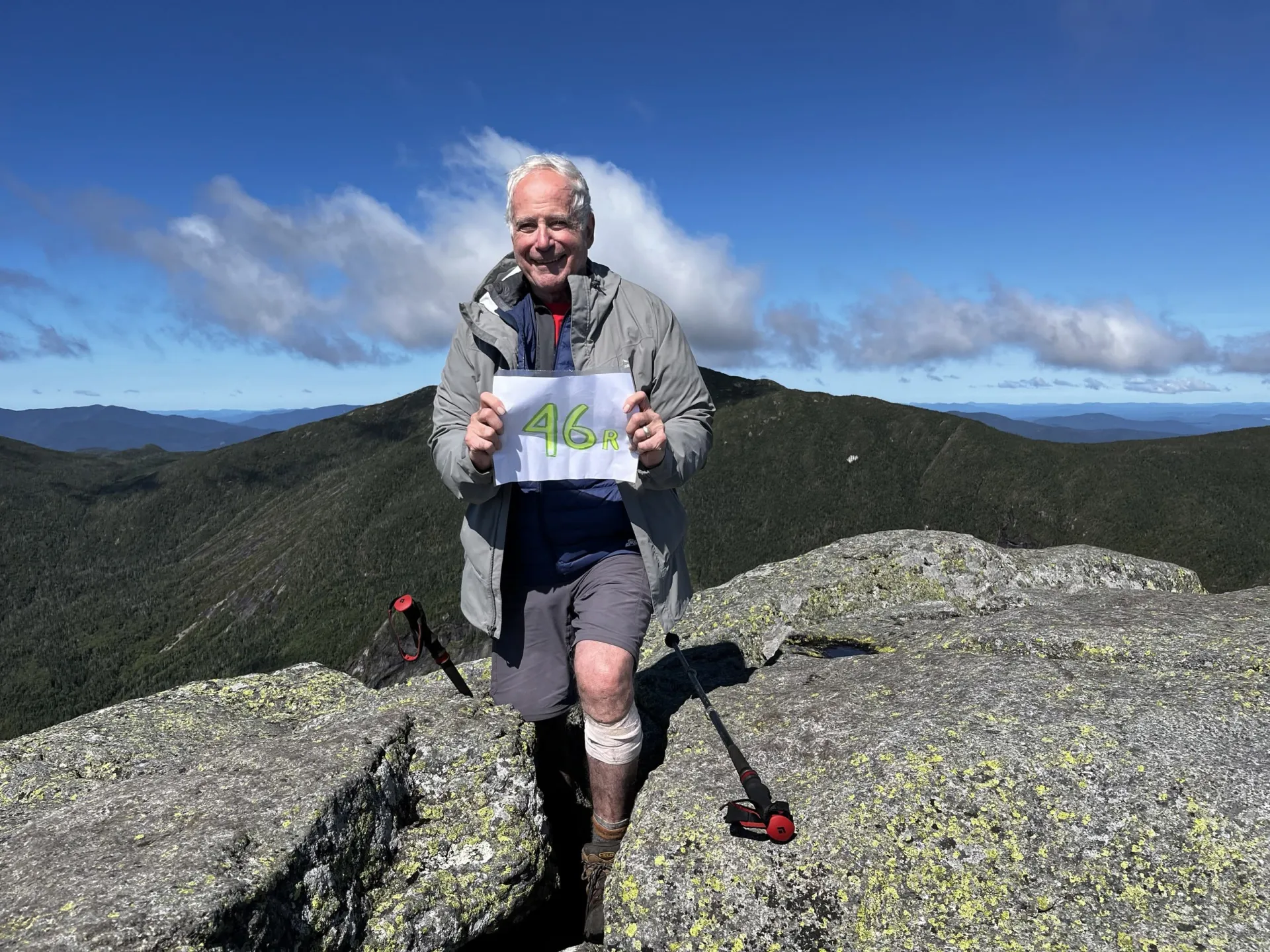 Older Gentleman stands atop a mountain peak holding a sign that reads '46er!'.