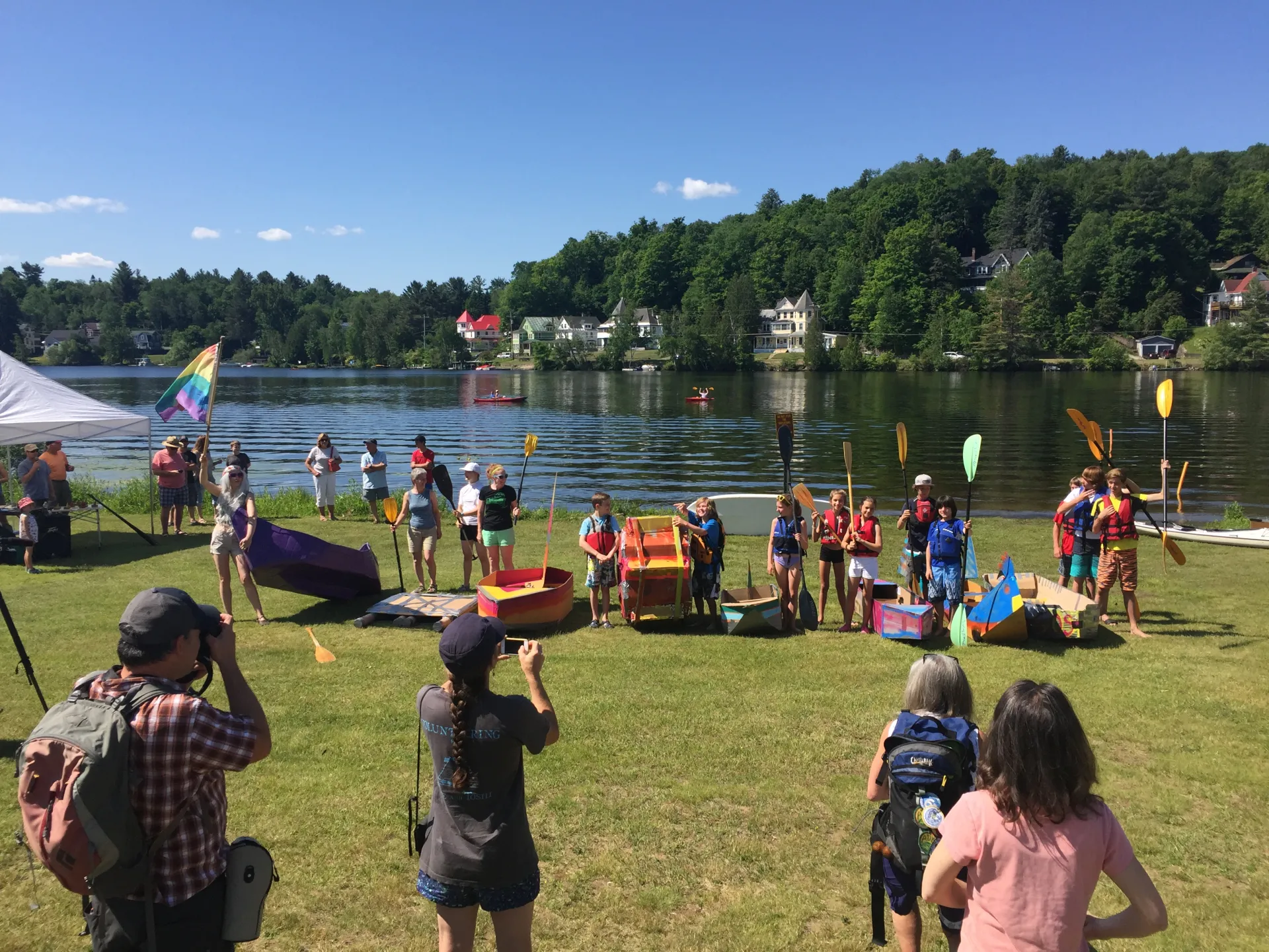group of people at the lake shore with cardboard boat materials and oars, getting ready to go in the lake 