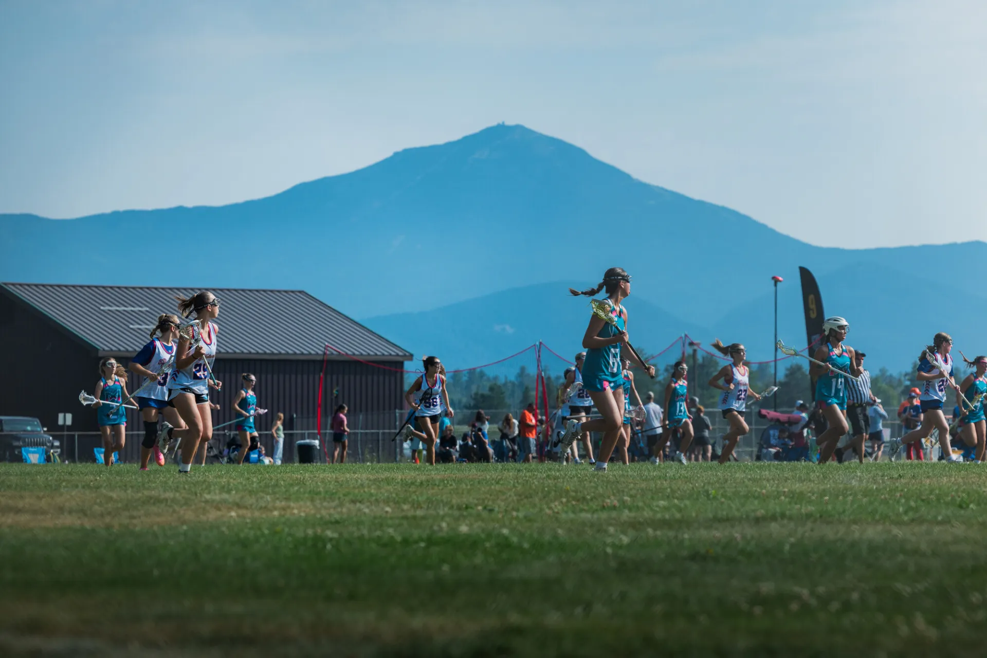 Female lacrosse players from two teams run down field with Whiteface Mountain in background