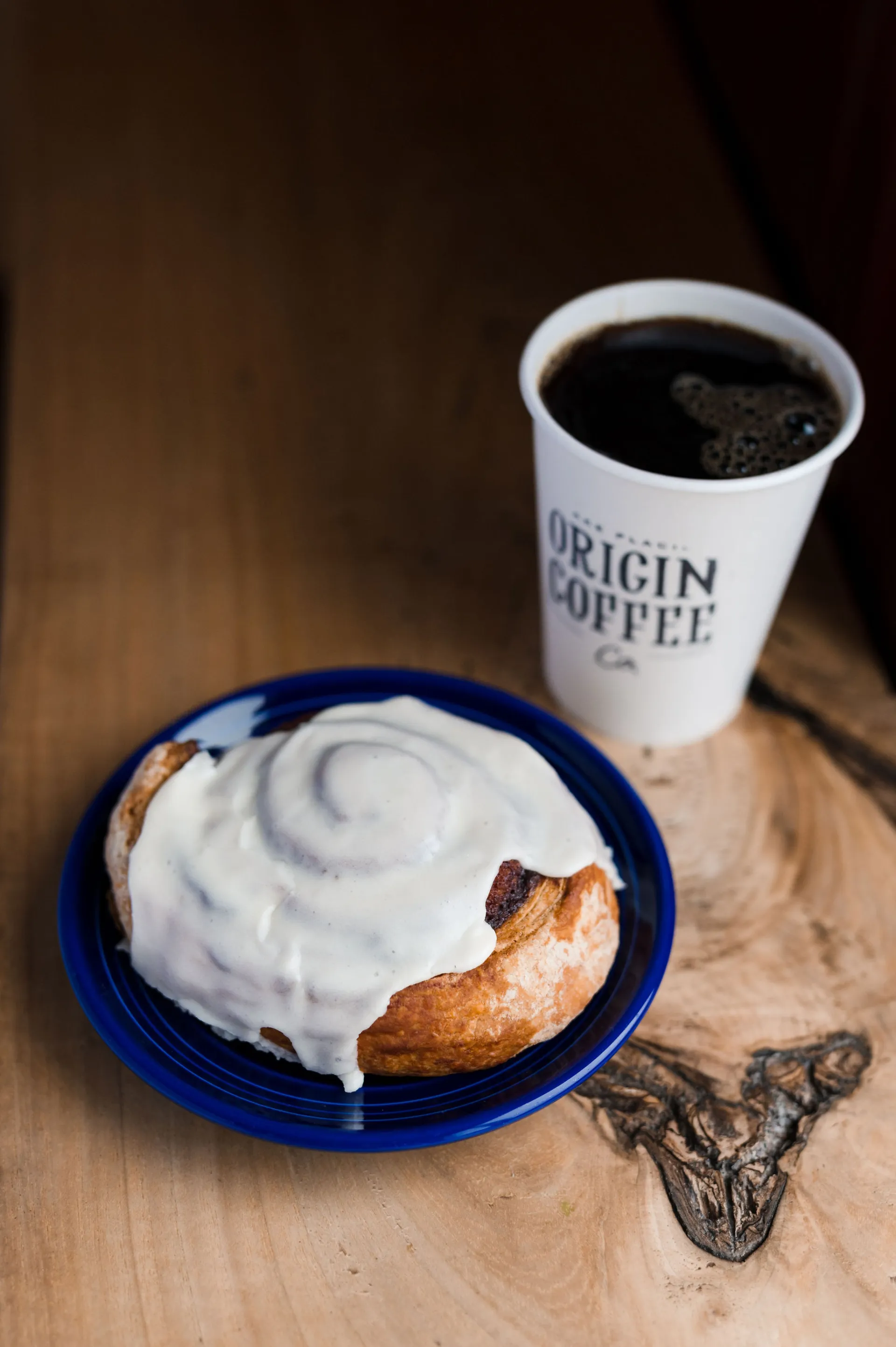 A large cinnamon bun, smothered in icing on a blue plate next to a black drip coffee in a branded Origin coffee cup.