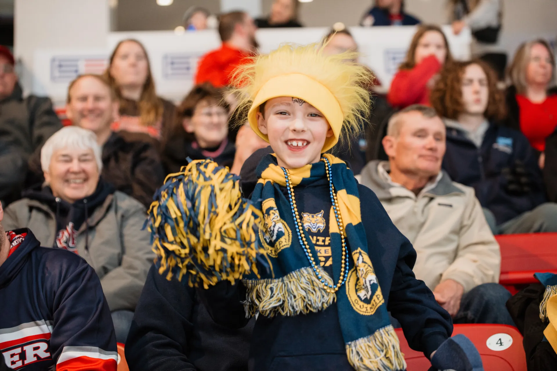 Child dressed in Quinnipiac hockey gear and accessories looks at camera and smiles while cheering in the stands.