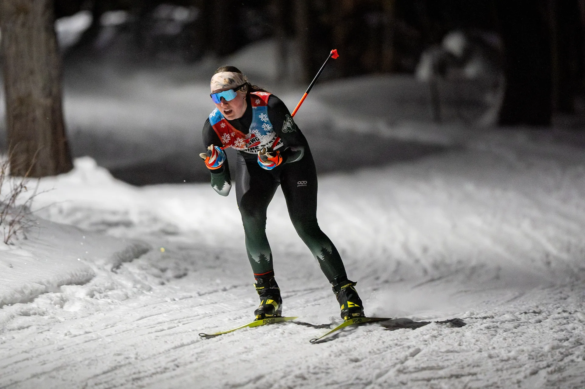 A cross-country skier in a racing suit and sunglasses glides along a snowy trail at night under bright artificial lighting.