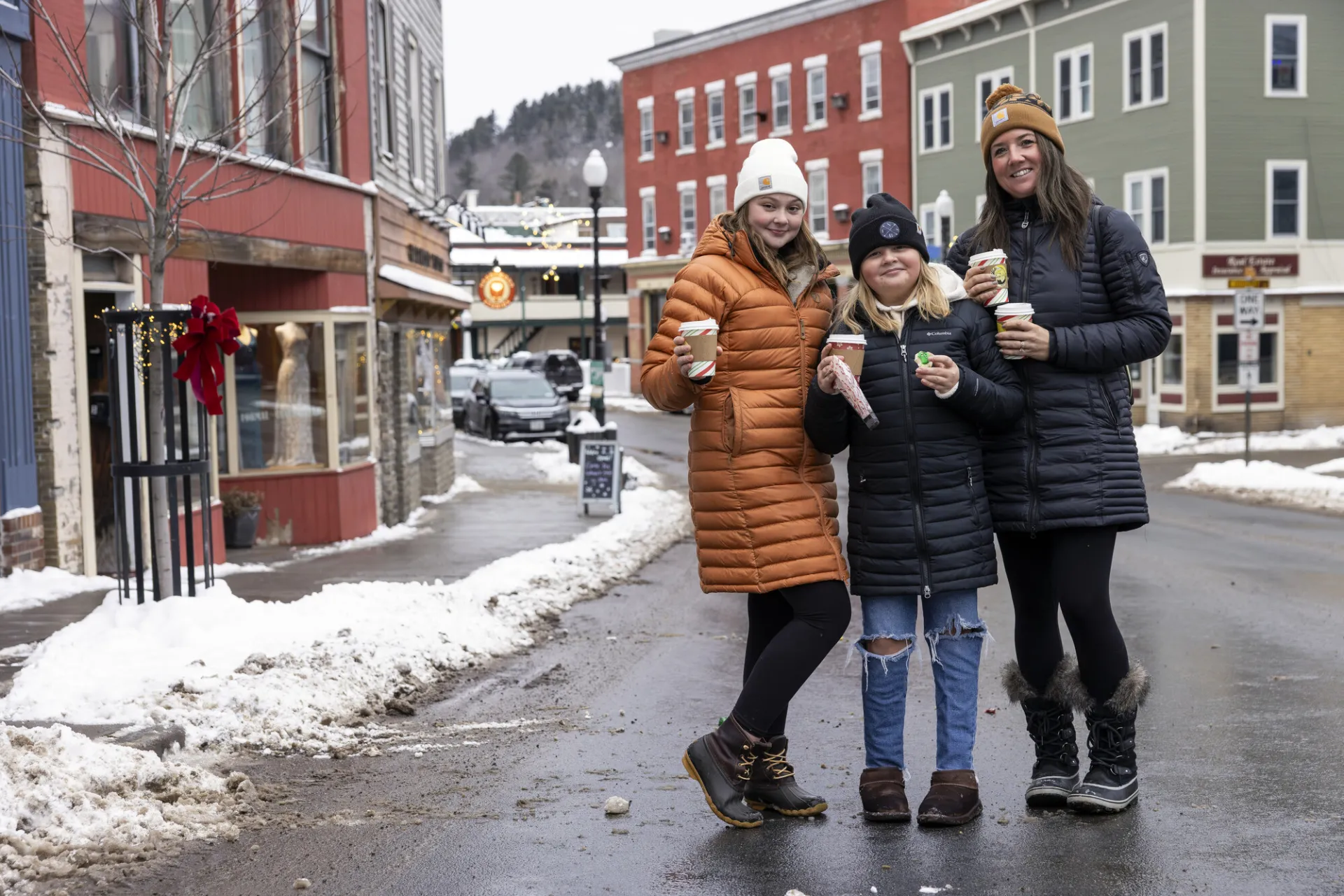 Three women standing in the middle of a winter village smile with hot cocoa in their hands. 