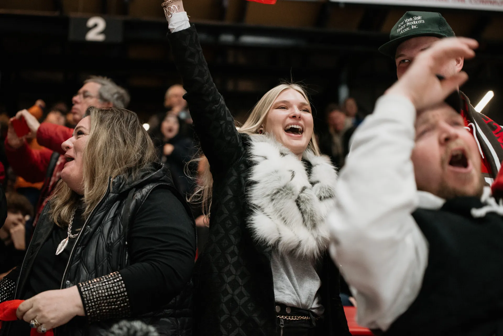 Woman dressed in black with furry white scarf holds arm above her head while cheering in the stands.