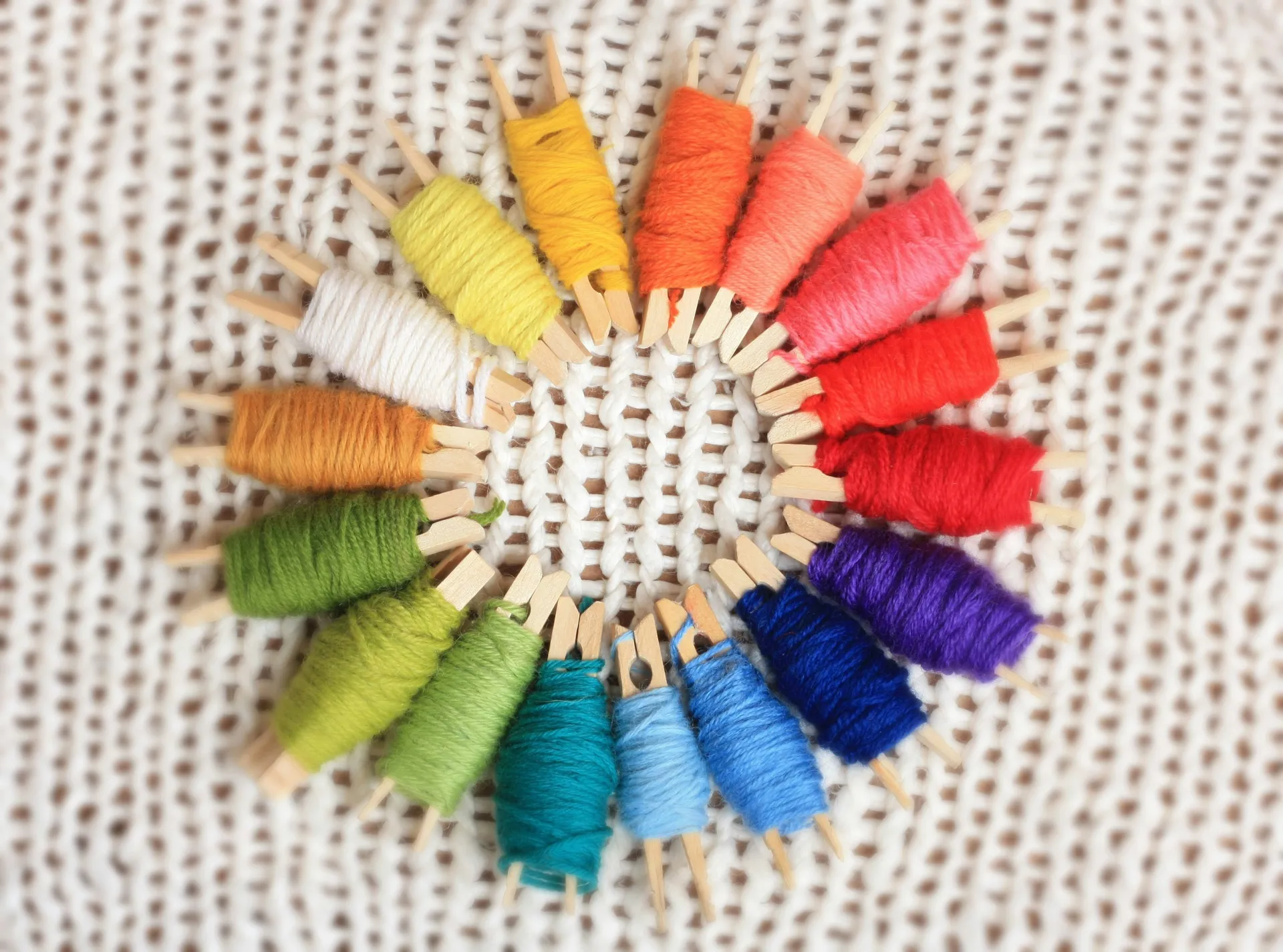 A view of colorful yarns wrapped around wooden clothespins, arranged in a rainbow wheel on a white knitted background.