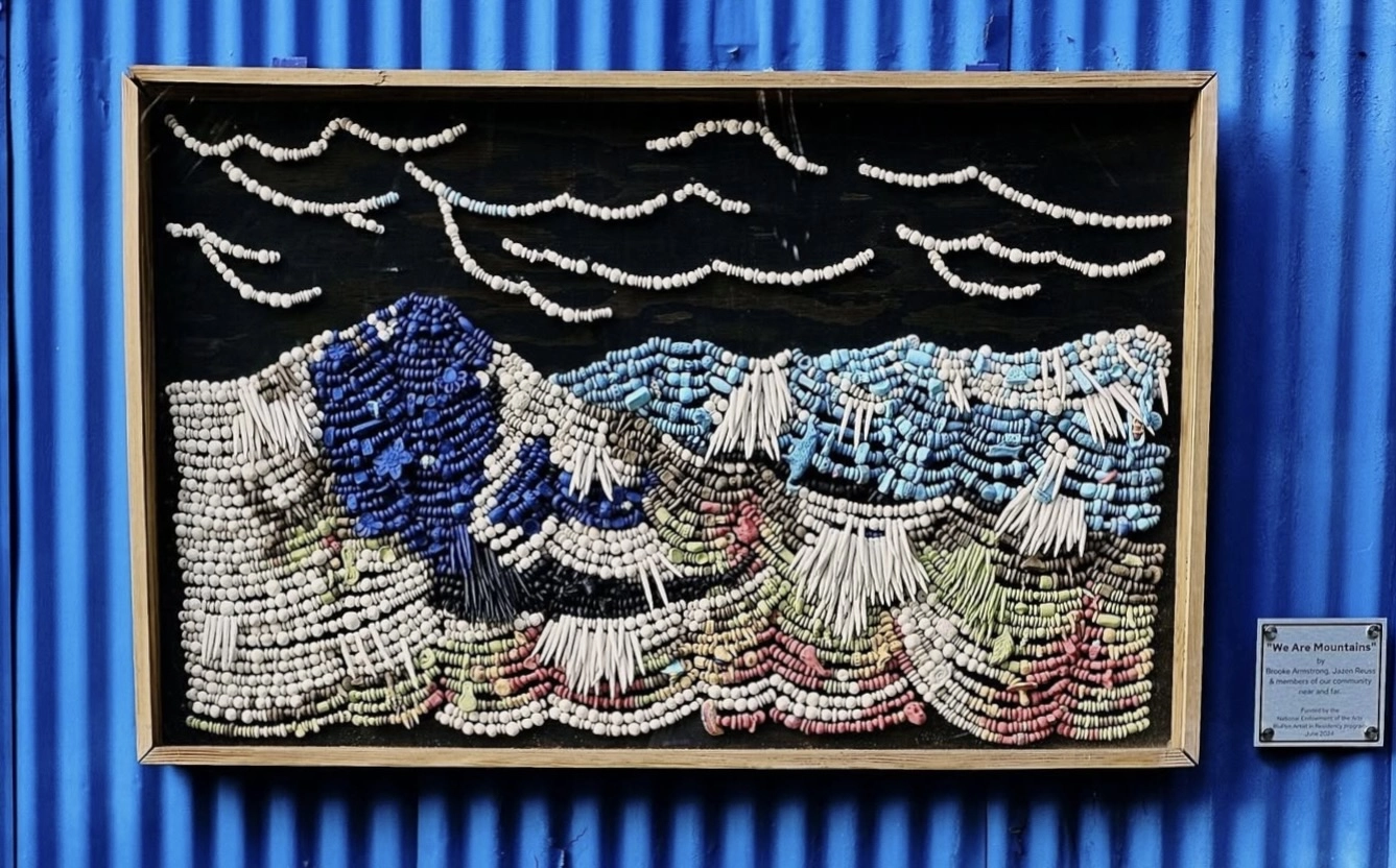 A framed artwork of a beaded landscape hangs on a corrugated blue building.