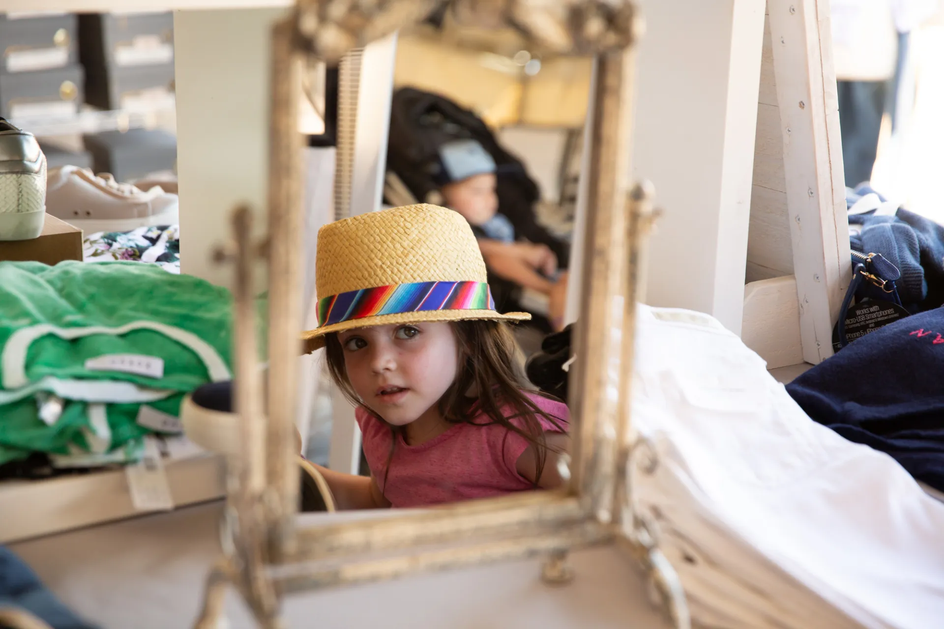 A little girl checks her self out in a mirror while wearing a straw hat with rainbow ribbon.