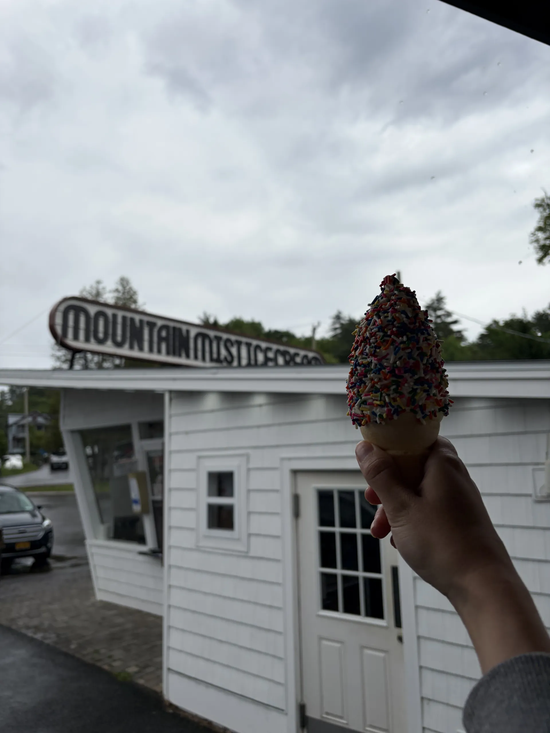 A hand holds up an ice cream cone in front of an ice cream stand.