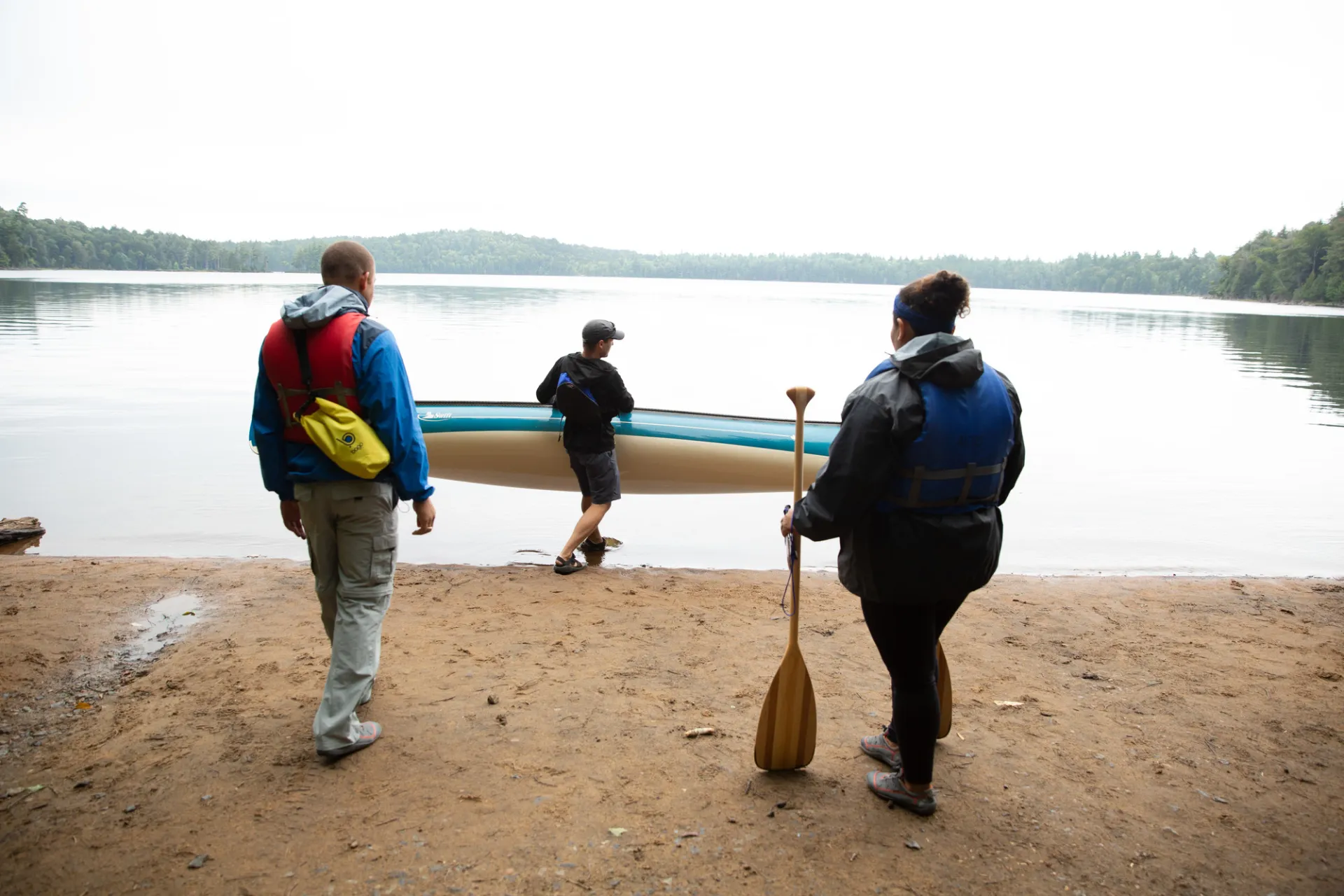 A man and woman watch as a man puts a canoe into a lake for them on a misty day.