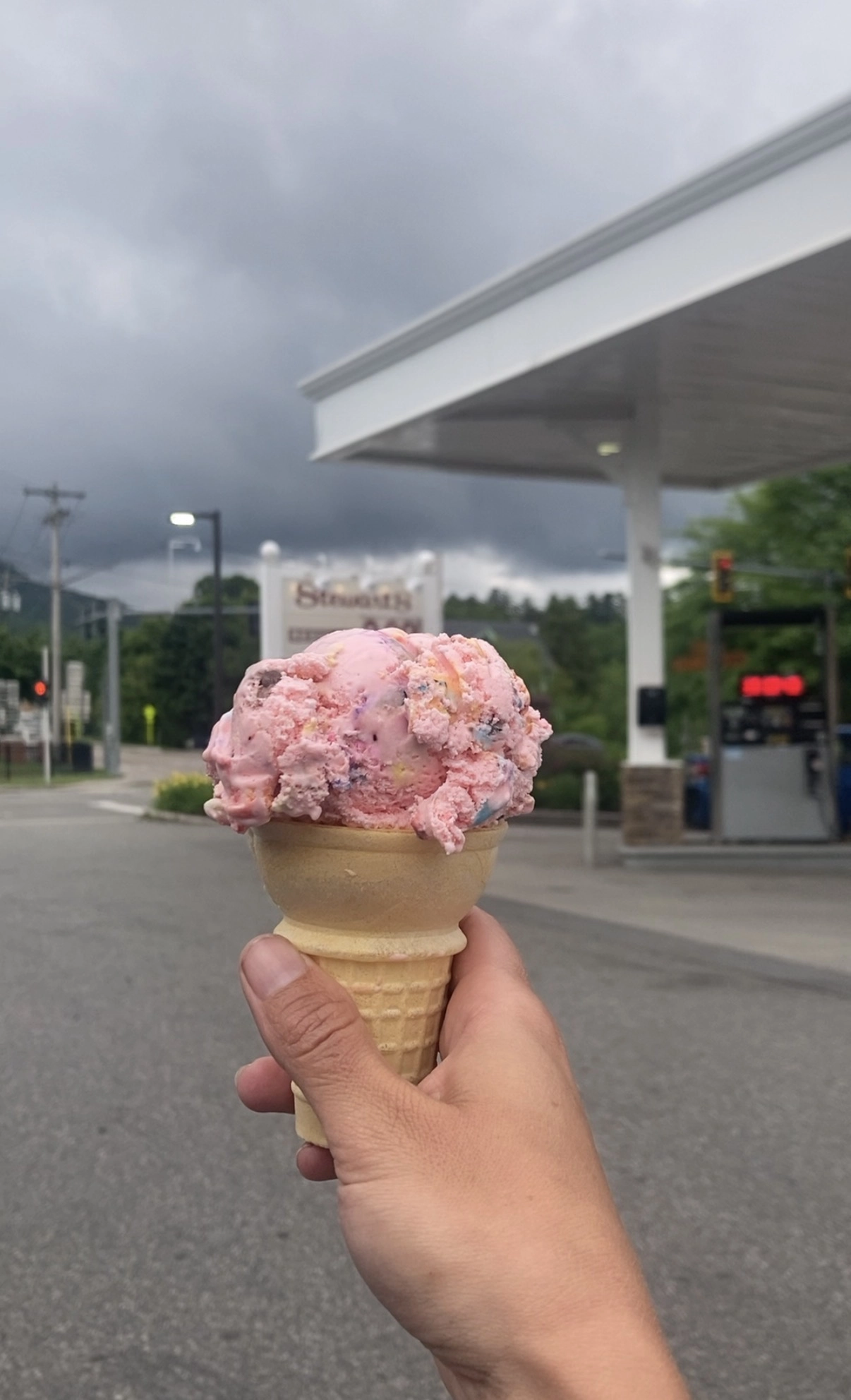 A hand holds up an ice cream cone in front of a gas station.