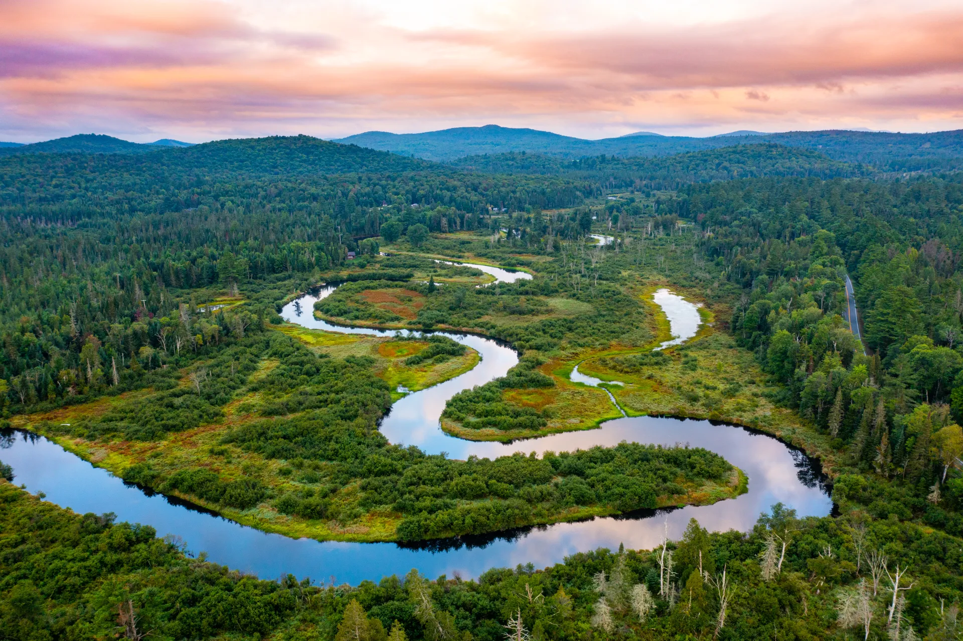 An aerial view of a calm river winding through a marshy wilderness. The sky is pink with sunset.