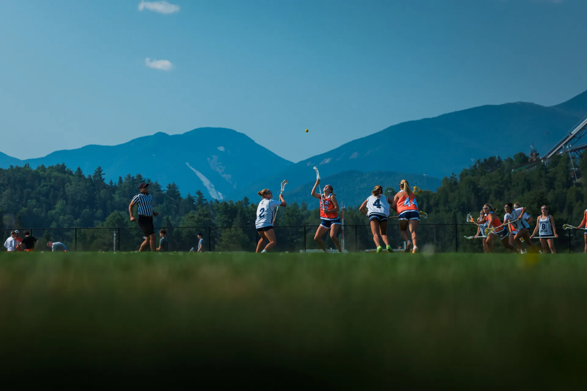 Two female lacrosse players look up at yellow lacrosse ball during play on field in front of Adirondack Mountain backdrop