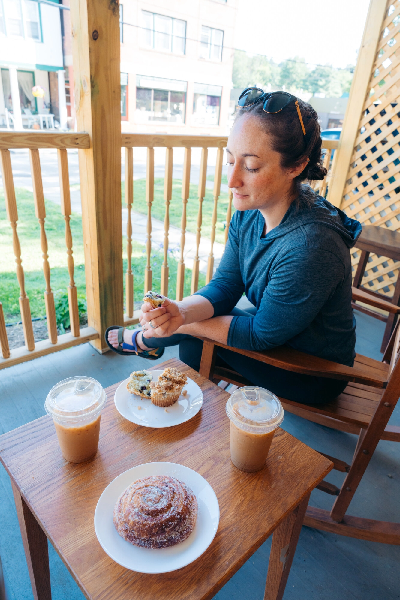 A woman sits in a rocking chair with a muffin and iced coffee on a small table next to her. 