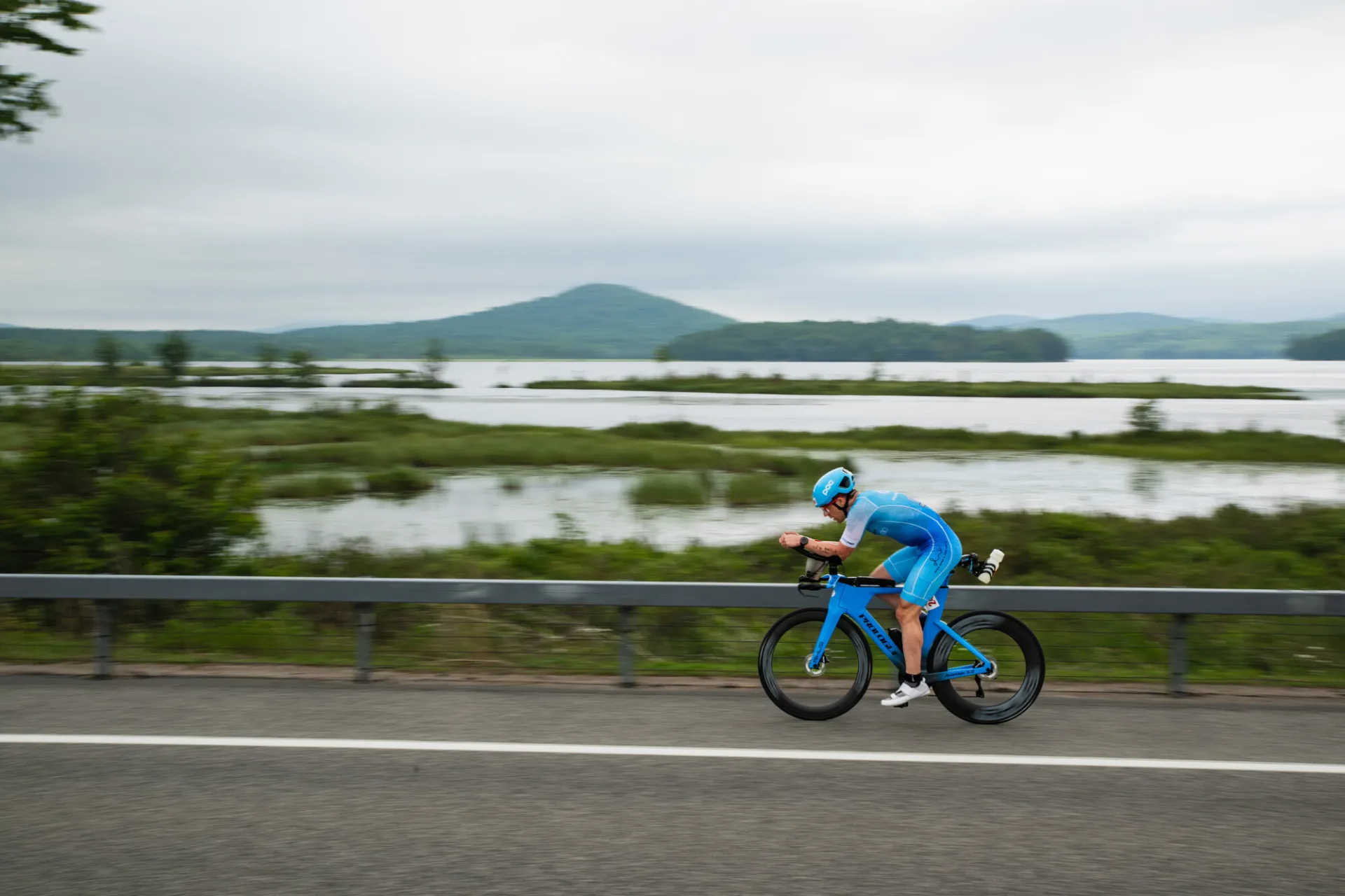 A triathlete in a bright blue speed suit and matching helmet rides a blue aerodynamic bike along a lakeside road.