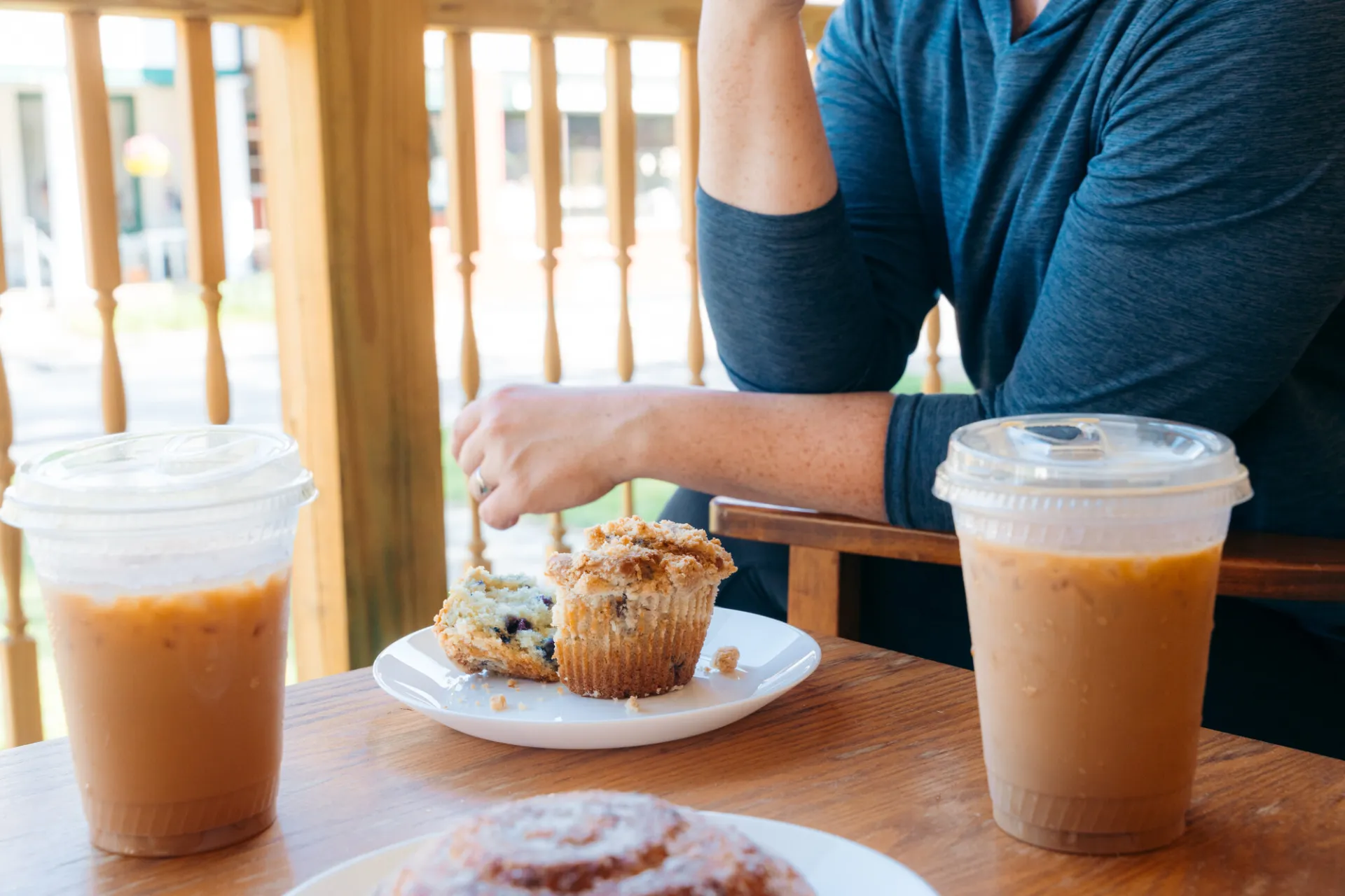A woman sits outside on a patio with a side table. On the table is an iced coffee, a muffin, and a cinnamon bun.