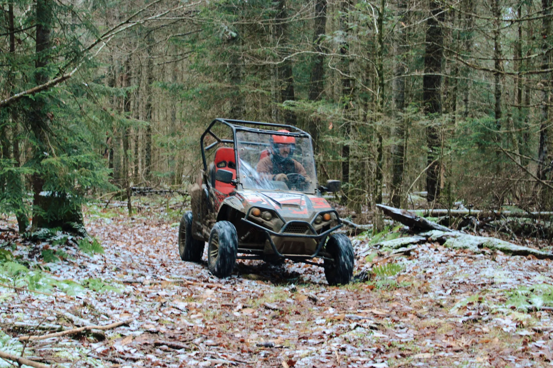 Early snow covers the fallen leaves as a man in a helmet drives a UTV on a path through the trees 